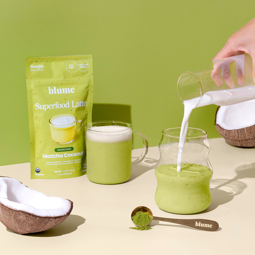 A package of blume Superfood Latte Matcha Coconut mix is displayed with a glass mug of green latte. A hand pours milk into a pitcher of green drink. A spoon with green powder and halved coconuts are nearby, set against a green background.
